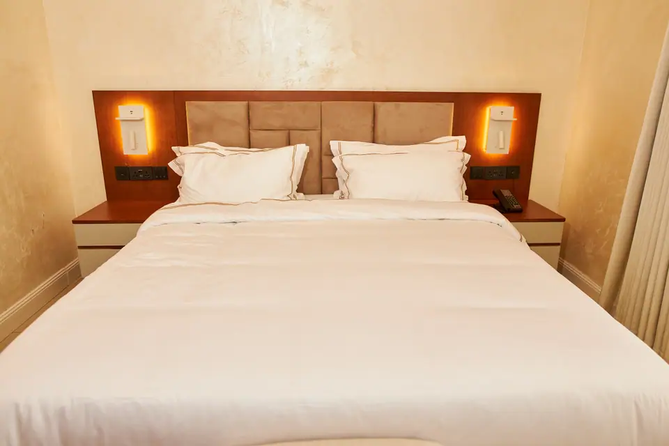 Deluxe room — king bed with padded headboard, gold-stitched white linens and warm amber wall sconces